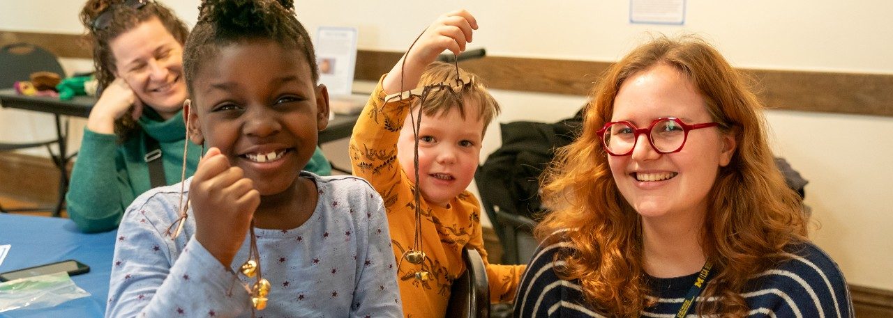 Two children and a woman smile at the camera at a Kelsey Family Day. The two kids hold up crafts made of bells and yarn.