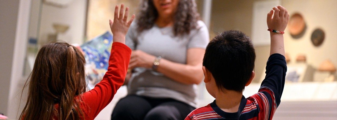 A woman reads to two children with their hands raised in the Kelsey Museum.