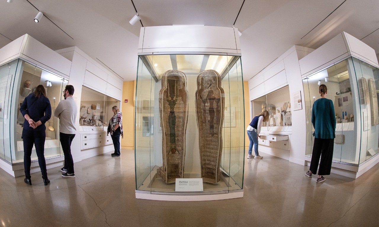 View of the Kelsey Museum’s Dynastic Egypt galleries, with the coffin of Djehutymose as the centerpiece. Visitors look at artifacts in display cases.