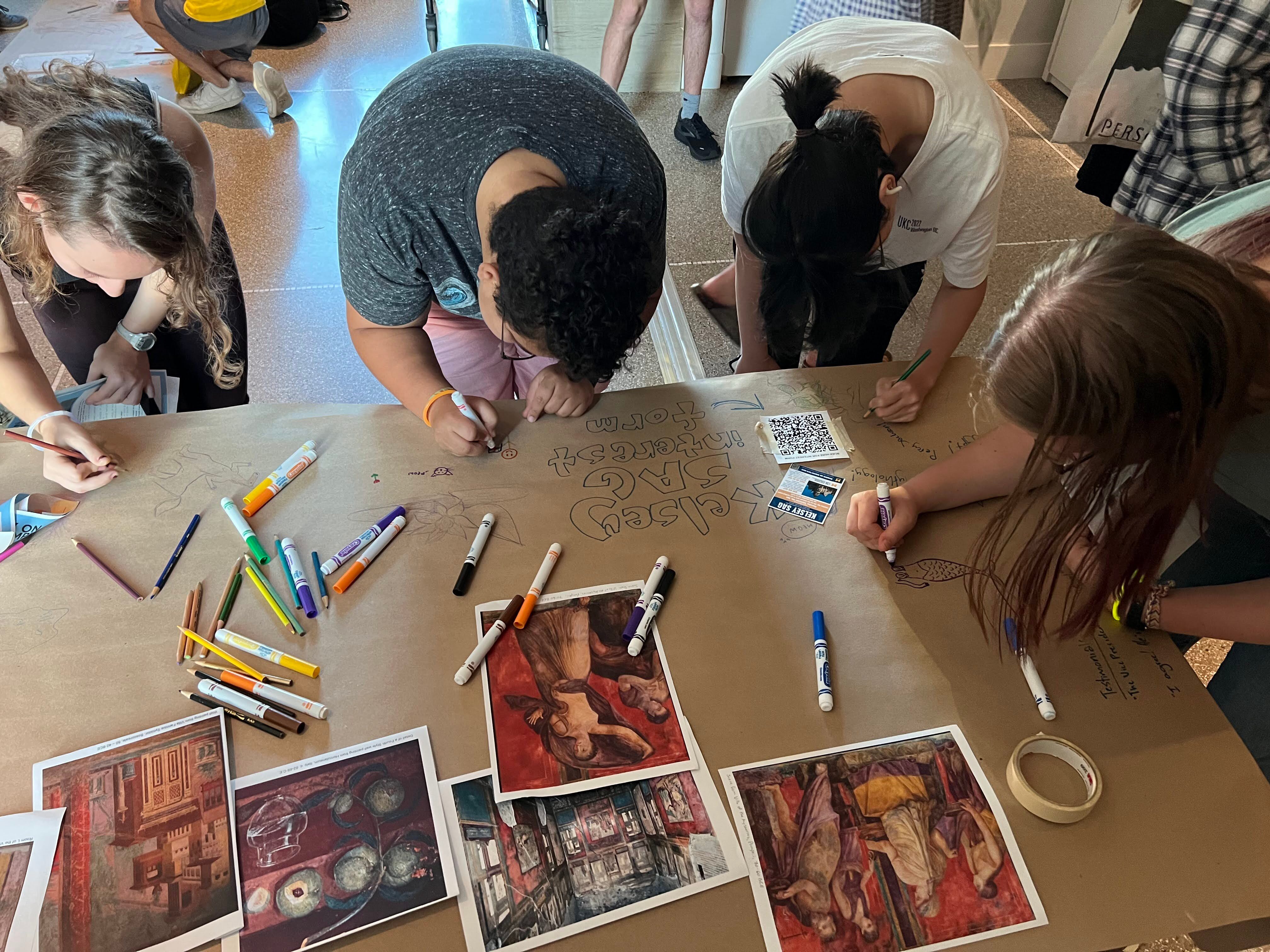 Four students use colored pencils and markers to draw on a brown paper tablecloth. Printed sheets of paper with Kelsey Museum artifacts and images rest on the table. 