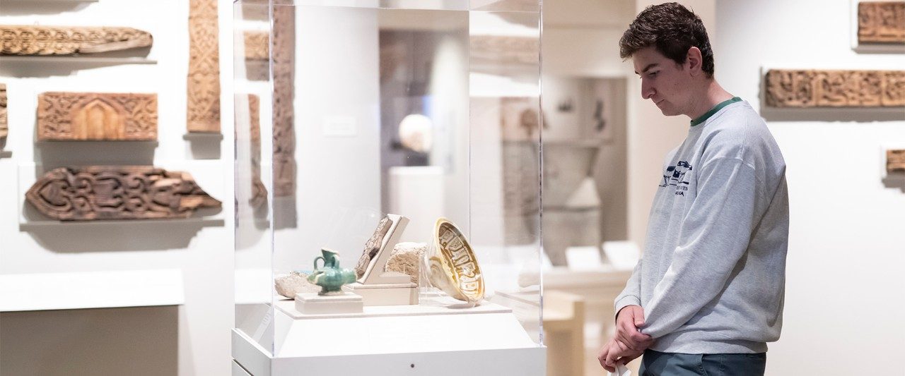 A group of college students in the Kelsey Museum’s Graeco-Roman Egyptian galleries. They face away from the camera, focused on a person out of view addressing them.