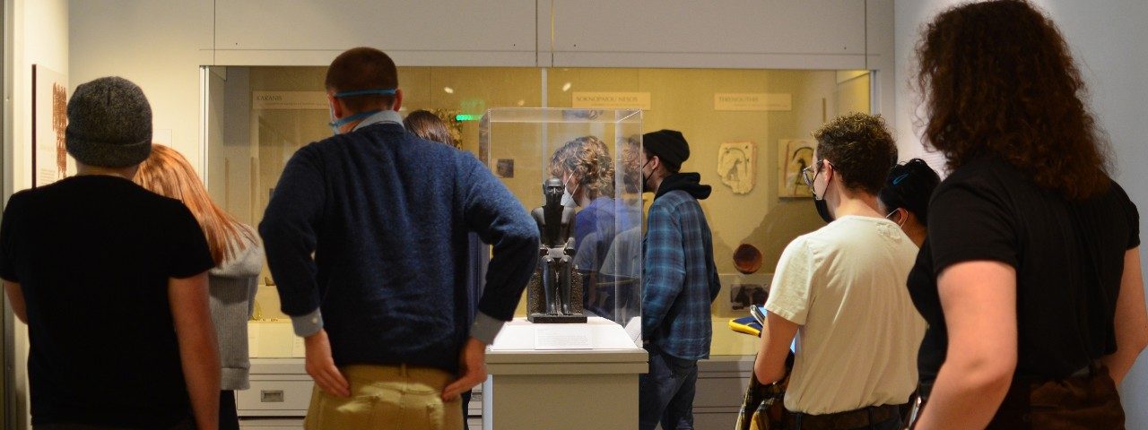 A group of college students in the Kelsey Museum’s Graeco-Roman Egyptian galleries. They face away from the camera, focused on a person out of view addressing them.