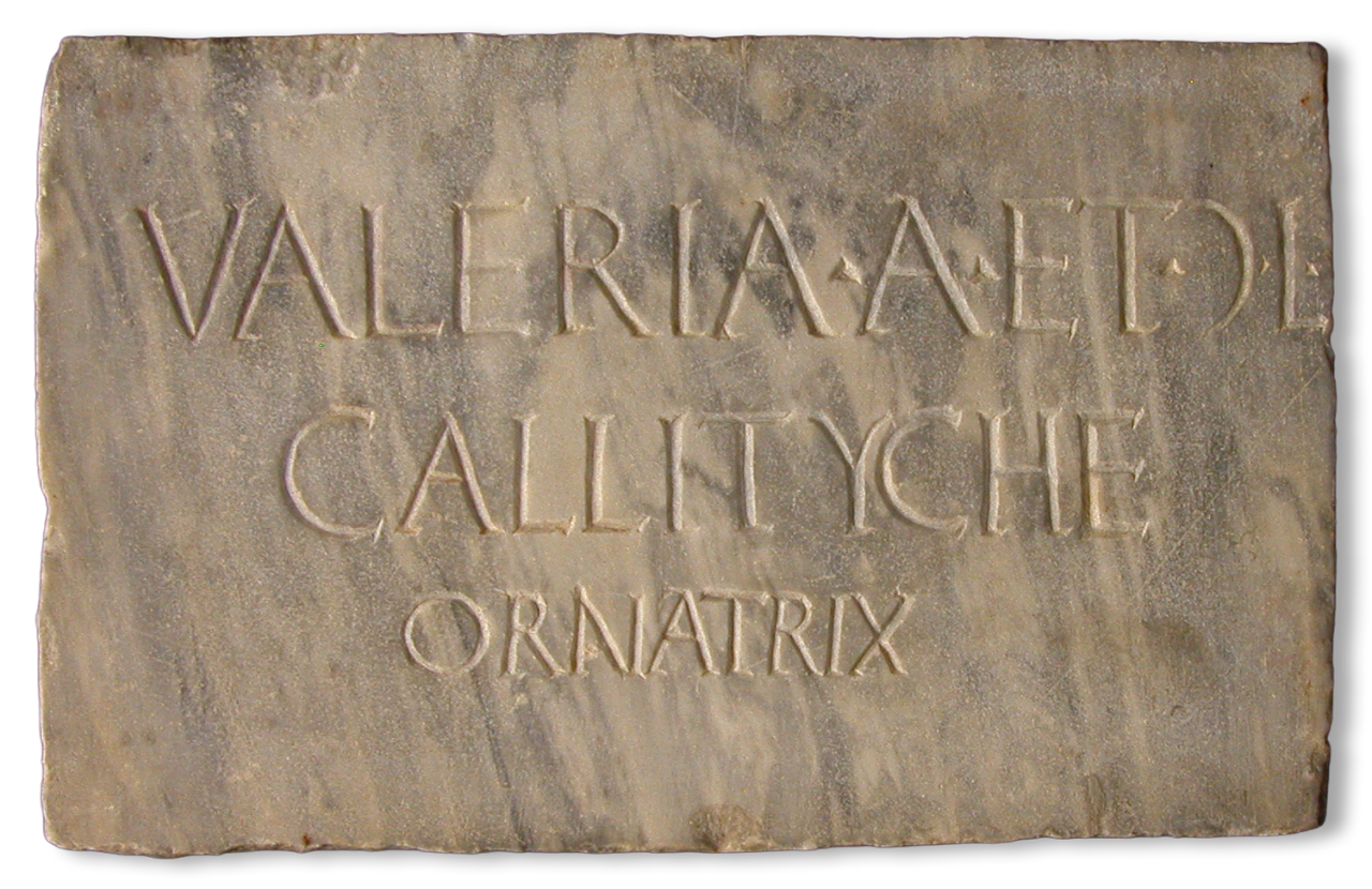 Light tan rectangular marble slab with a Latin inscription reading “Valeria A(uli) et ((C))(aiae) liberta / Callityche / ornatrix.” Red pigment is faintly visible in some of the letters.