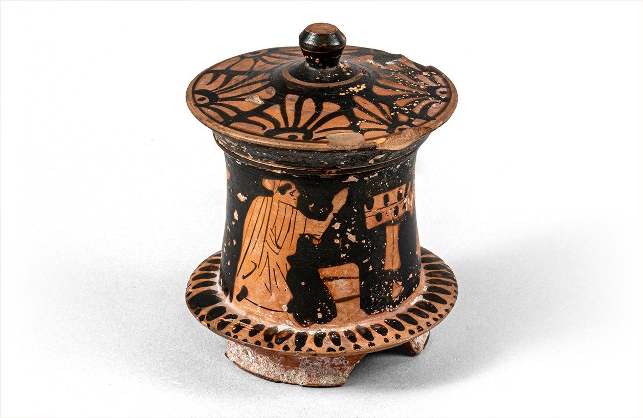 A red-figure jar with a flared base and a fitted lid decorated with flower patterns. The surface of the jar shows a woman looking at herself in a mirror.