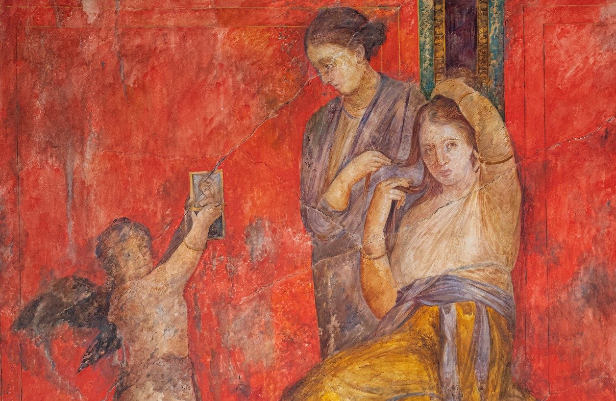 A vibrantly painted watercolor depicting a scene from the Pompeiian Villa of the Mysteries. In it, a winged boy holds up an image for an ornatrix, who style’s a seated woman’s hair.