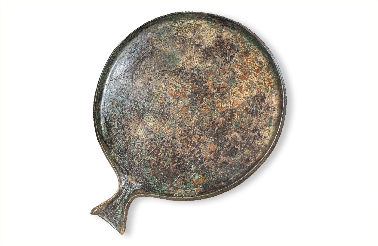 A round bronze mirror with a small handle. An etched image is faintly visible on its corroded surface.