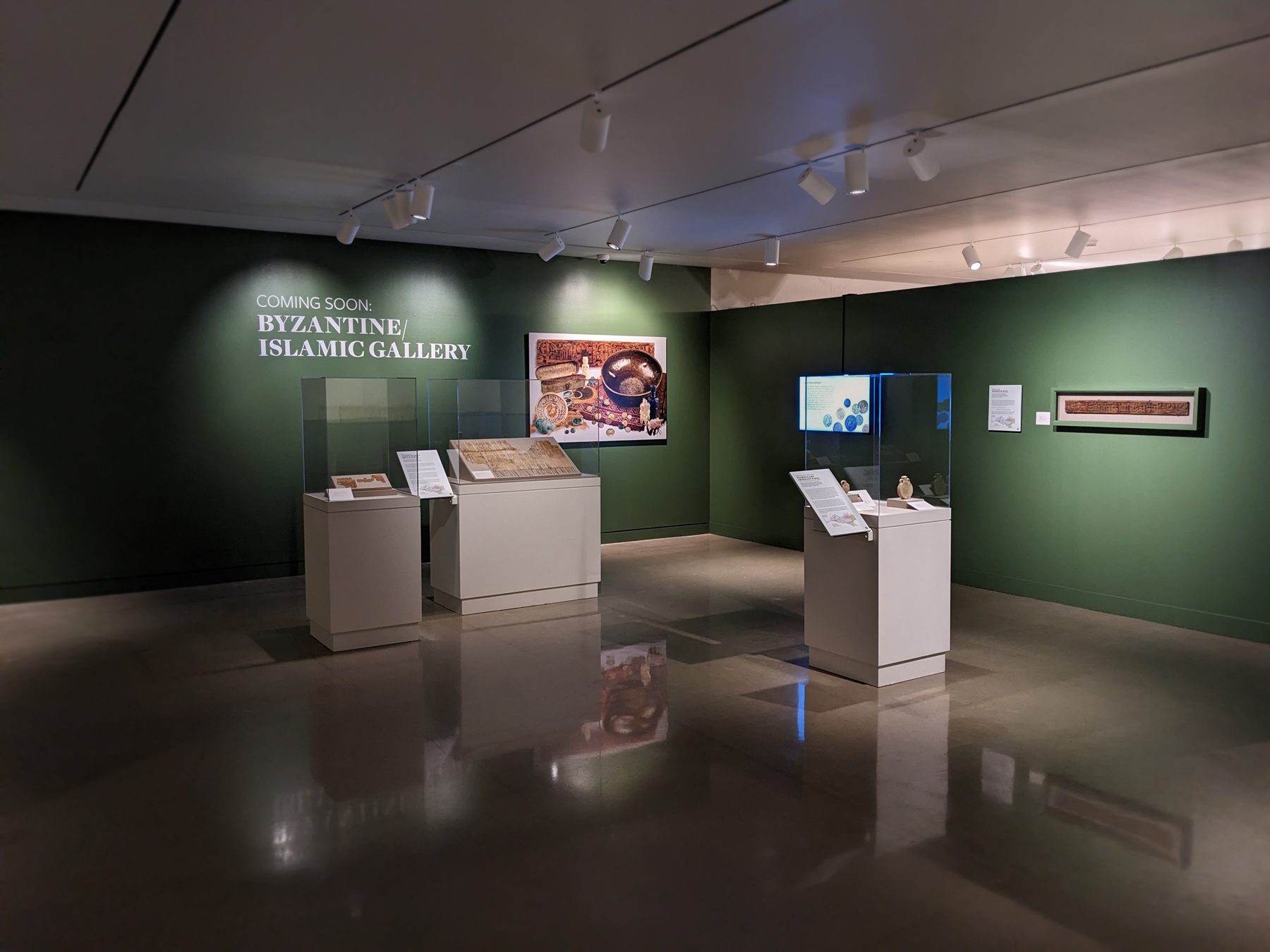 Museum gallery space with three vitrines containing textiles and ceramic artifacts. The walls are painted green and hold a photo of Byzantine and Islamic artifacts, a TV screen with a slideshow, and a mounted wooden door lintel. Text on the wall reads, “Coming Soon: Byzantine/Islamic Gallery.”