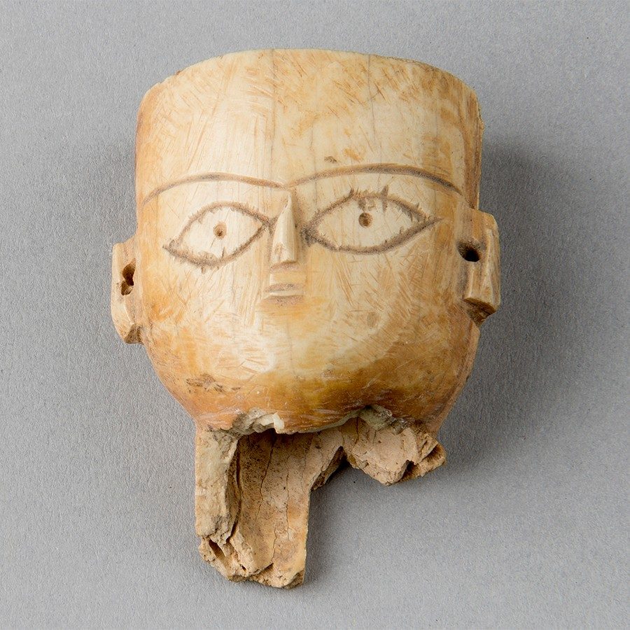 Tan-colored bone figurine of a head with a simply rendered carved face. The head has holes in its ears, and the bone is broken off at the neck.