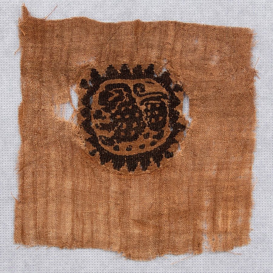 Tan square of fabric with a roundel at its center featuring a red, spotted lion encircled by a red sunburst.