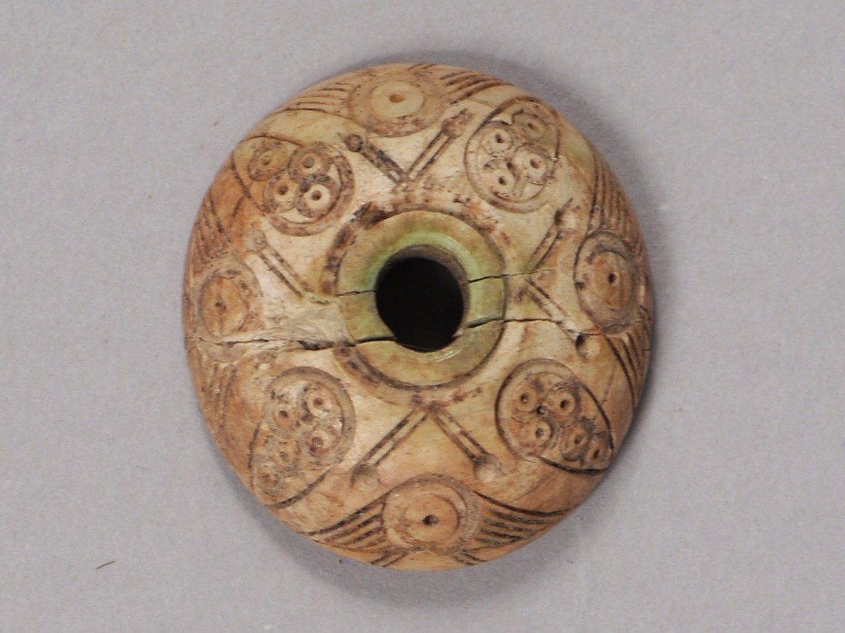 Small round object made out of bone with a hole bored through its center. The surface of the spindle whorl is covered with geometric patterns, including four carvings resembling skulls.