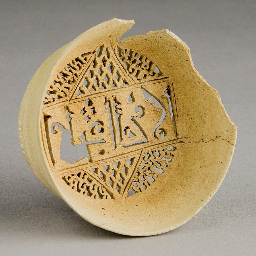 Buff-colored clay water filter with decorative punctures at its bottom. Across the top and bottom holes of various shapes, while enclosed within a central rectangle are punctures resembling Arabic script.