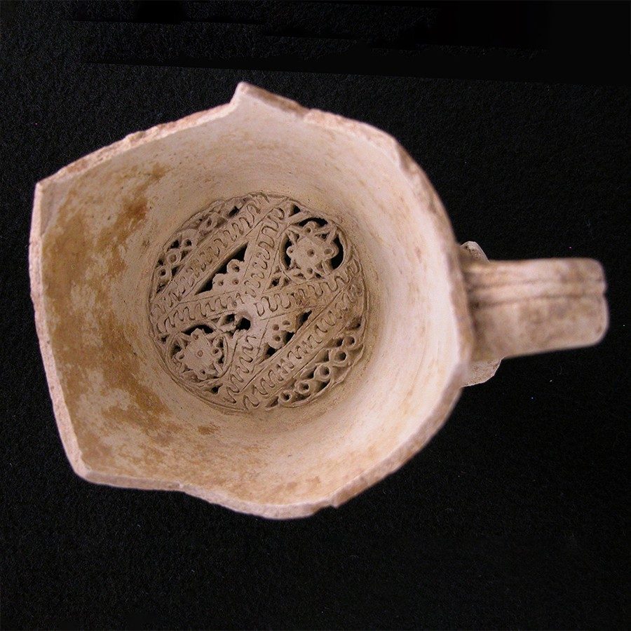 Buff-colored clay water filter with a geometric shapes radiating out from the center. The vessel also has a small handle.