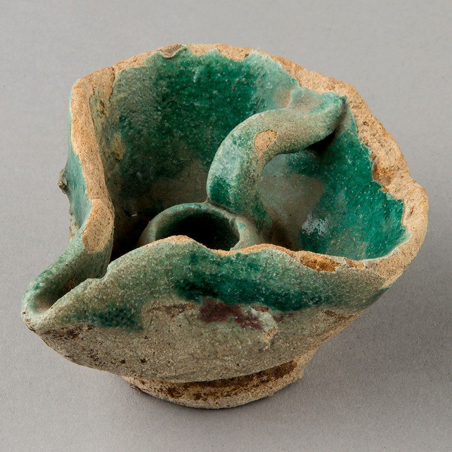 Clay oil lamp painted blue-green on a small, round base. A small handle sits on the interior of the object, near the wick hole. The oil chamber is irregularly shaped, pinched at one end.