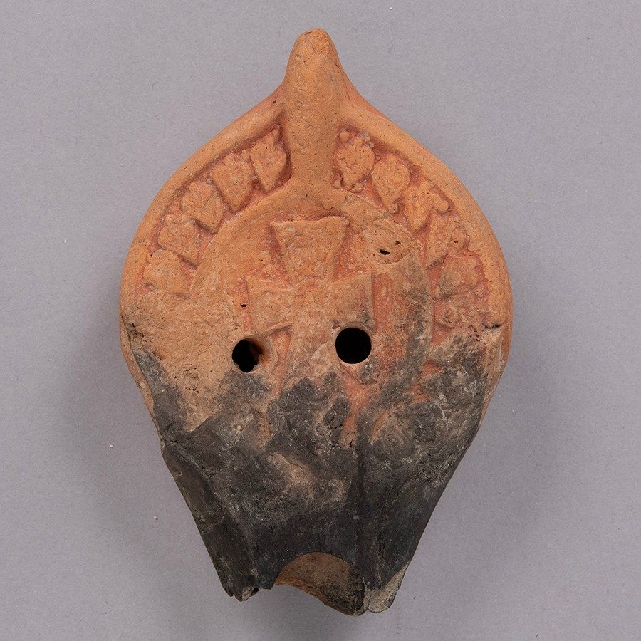 Red-orange terracotta oil lamp with a broken, blackened nose. The surface of the object has impressions of a Byzantine cross and a row of decorative leaves.