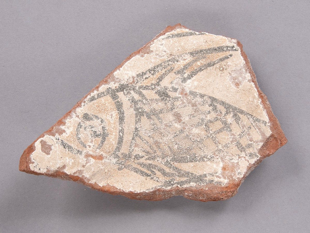Piece of ceramic with a whiteish cast painted with a fish with crisscrossing scales.