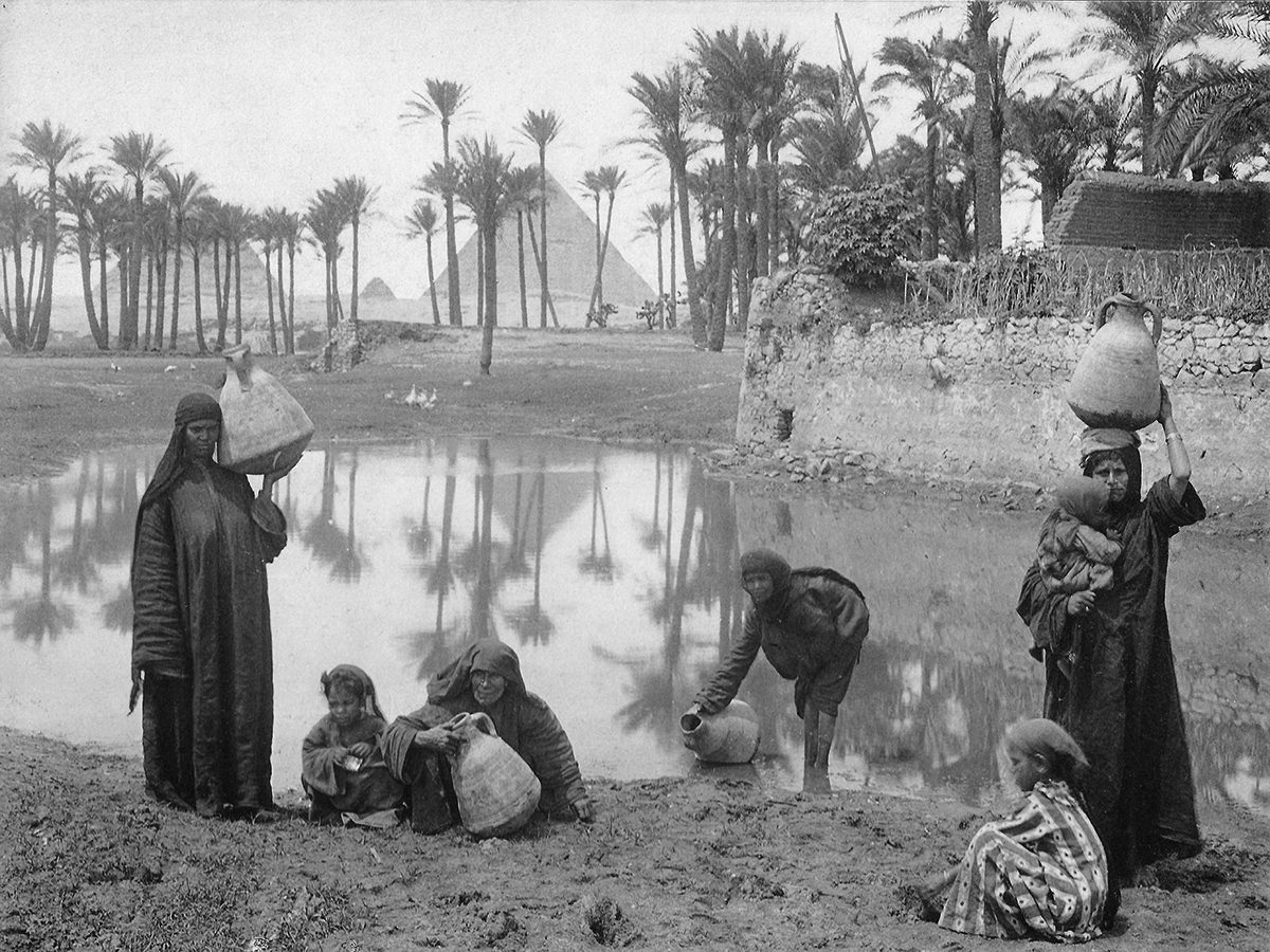 Archival photo showing a group of women and children filling large, two-handled vessels with water. In the background, palm trees and the Giza pyramids are visible.