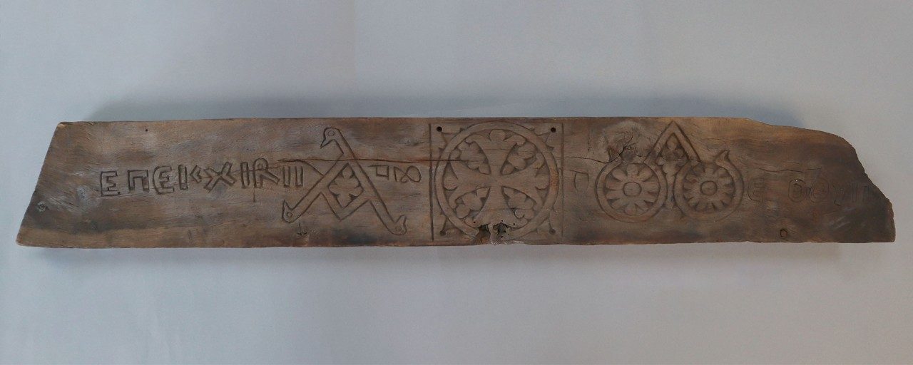 Long, rectangular wooden relief panel carved with an inscription and a stylized alpha, Greek cross, and omega.