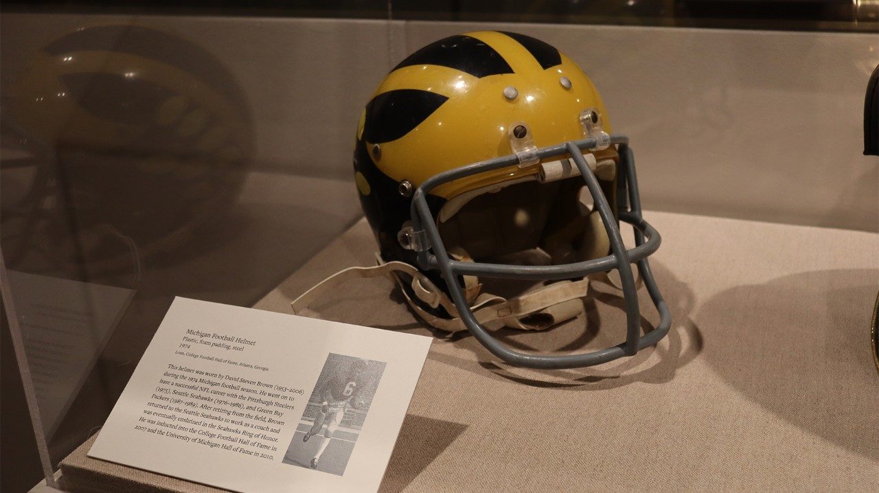 Blue and gold football helmet with a gray face guard, on display in a museum case.