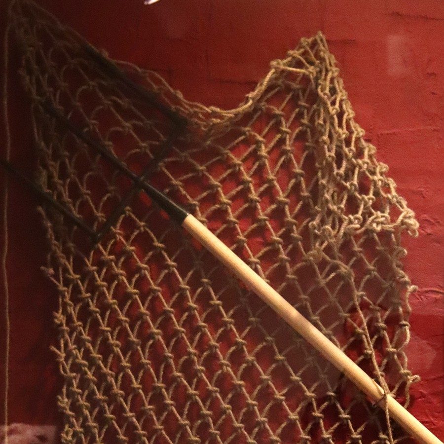 Steel trident with a wooden handle hung in front of a net on a red wall.
