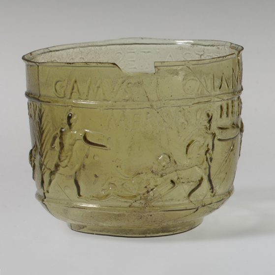 Green glass cup with a scene of gladiators fighting. Latin inscriptions line the top of the cup, near the rim. 