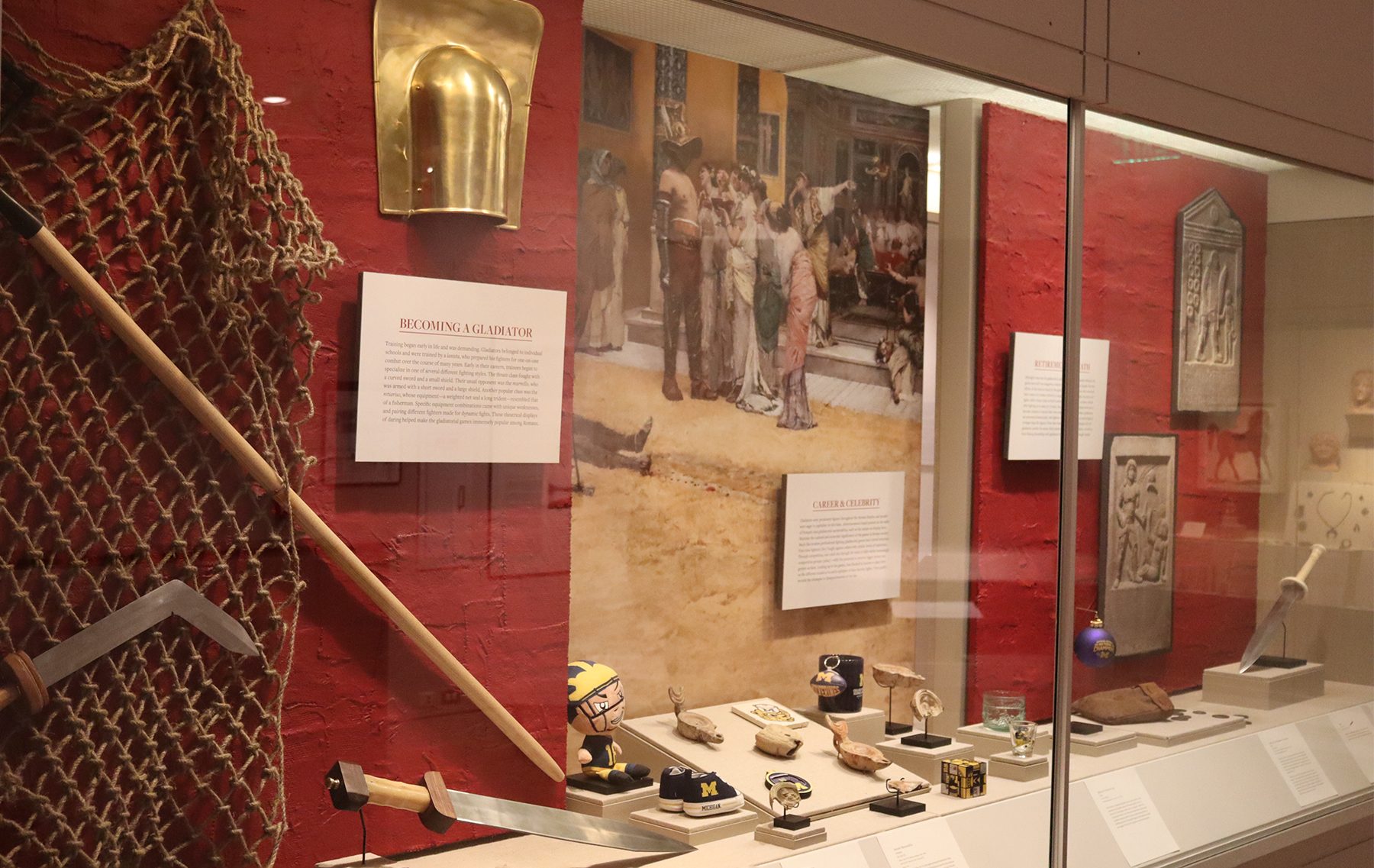 Museum case with replica artifacts, ancient oil lamps, modern University of Michigan memorabilia, a print of an oil painting depicting a gladiator surrounded by admirers, and photos of Roman tombstones.