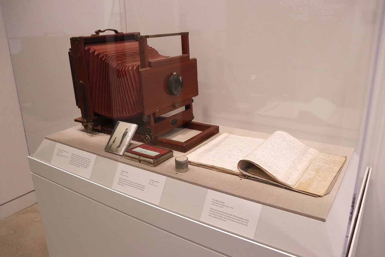 Museum case displaying, left to right, a large-format camera, a glass plate, a box of additional glass plates, a film canister, and a bound collection of handwritten letters.