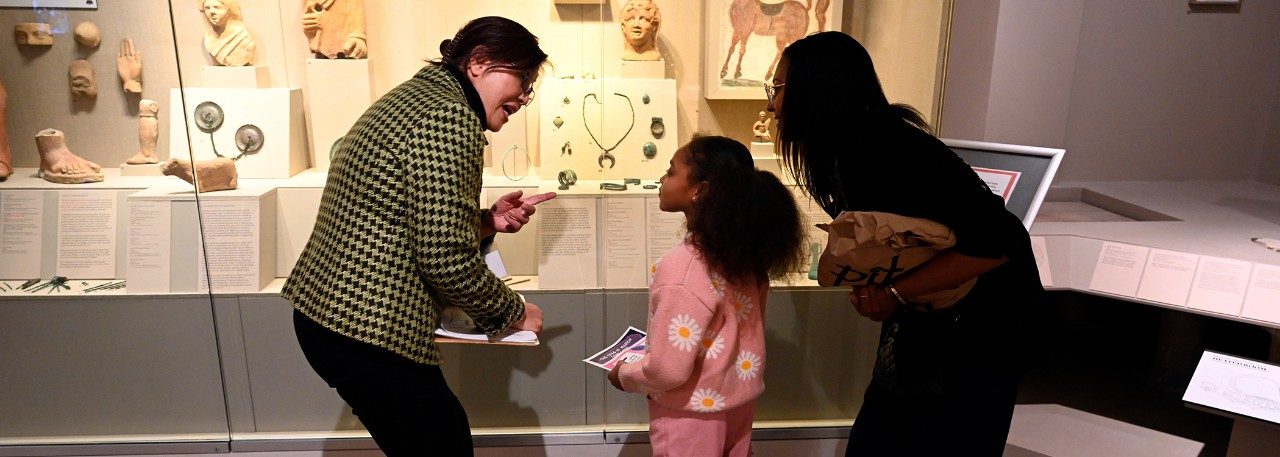 A docent stands with two visitors—a mother and daughter—in the Kelsey Museum’s Roman gallery.