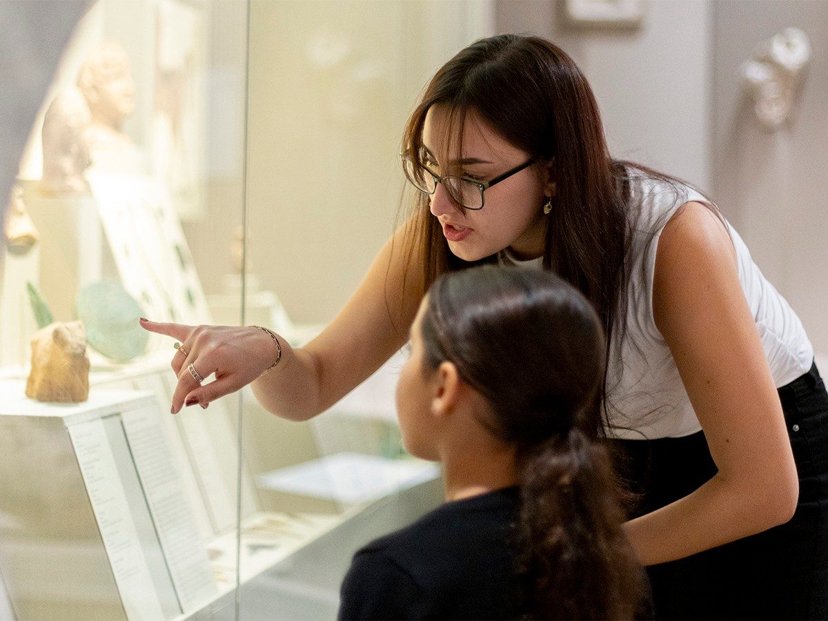 Undergraduate student Leyla Dumke talks to a child while pointing to an object in a museum case.