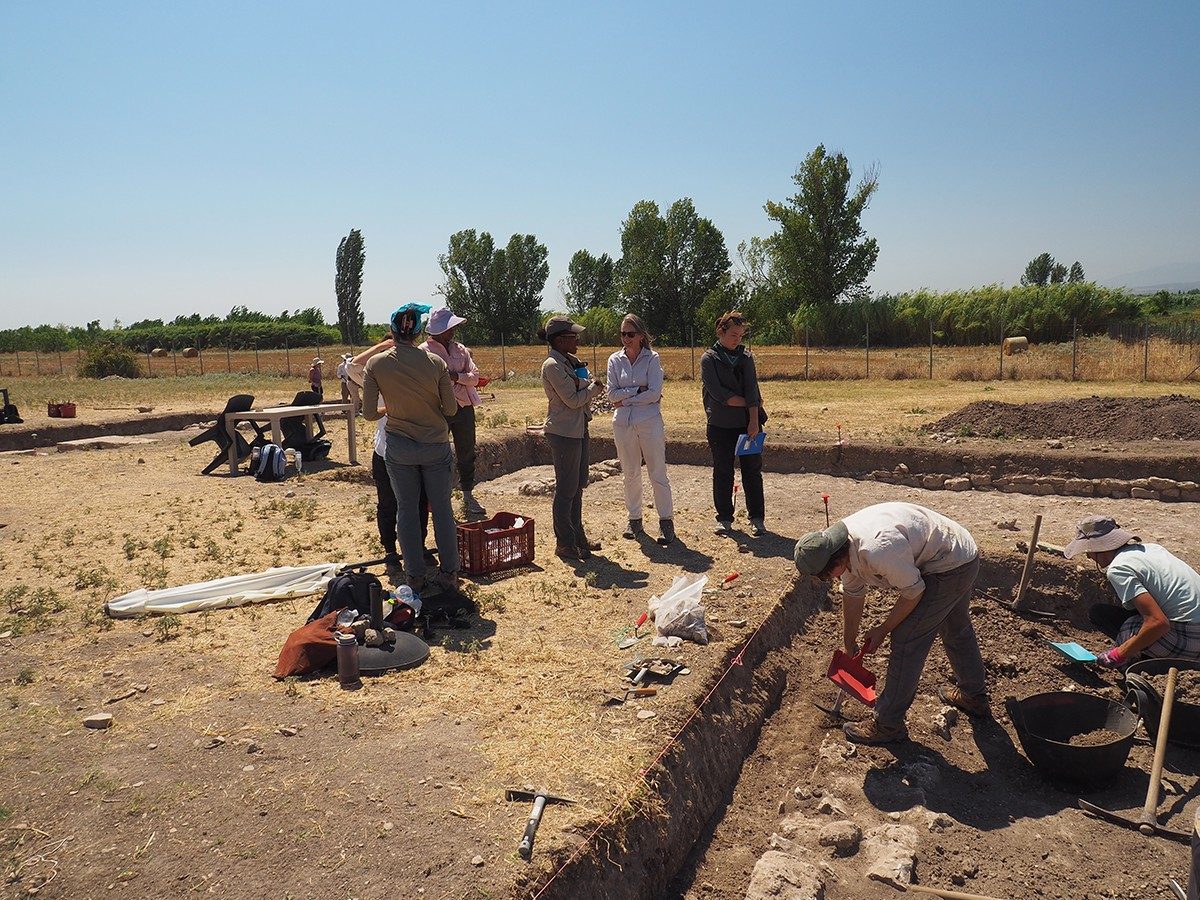 View of the archaeological site of Pella. A group of six people stands around a trench, in which two individuals excavate using pickaxes and dustpans.