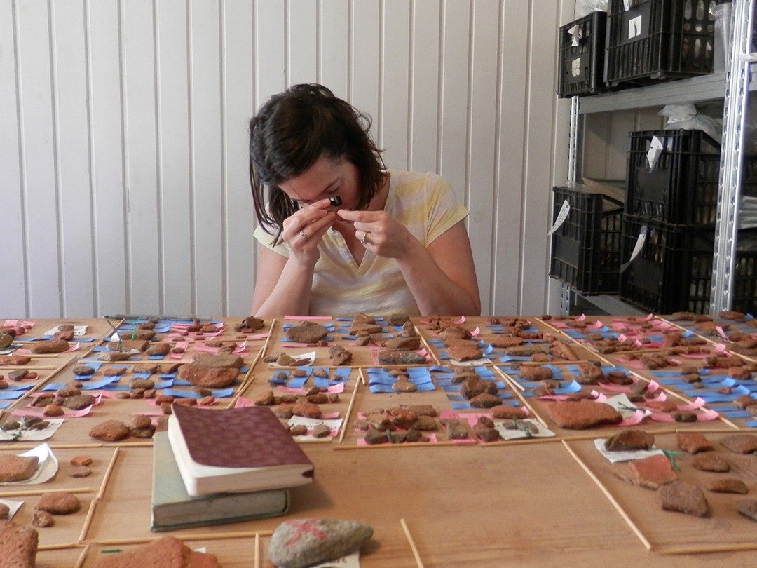 Natalie Abell uses a handheld magnifying device to examine a piece of pottery at a table covered in fragments.