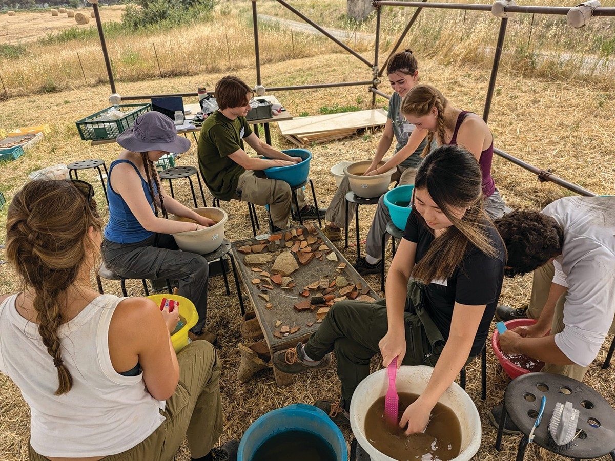 A group of seven people sits around a low table covered with pottery fragments. They each have a bucket full of water and a brush, which they use to clean dirt off the sherds.