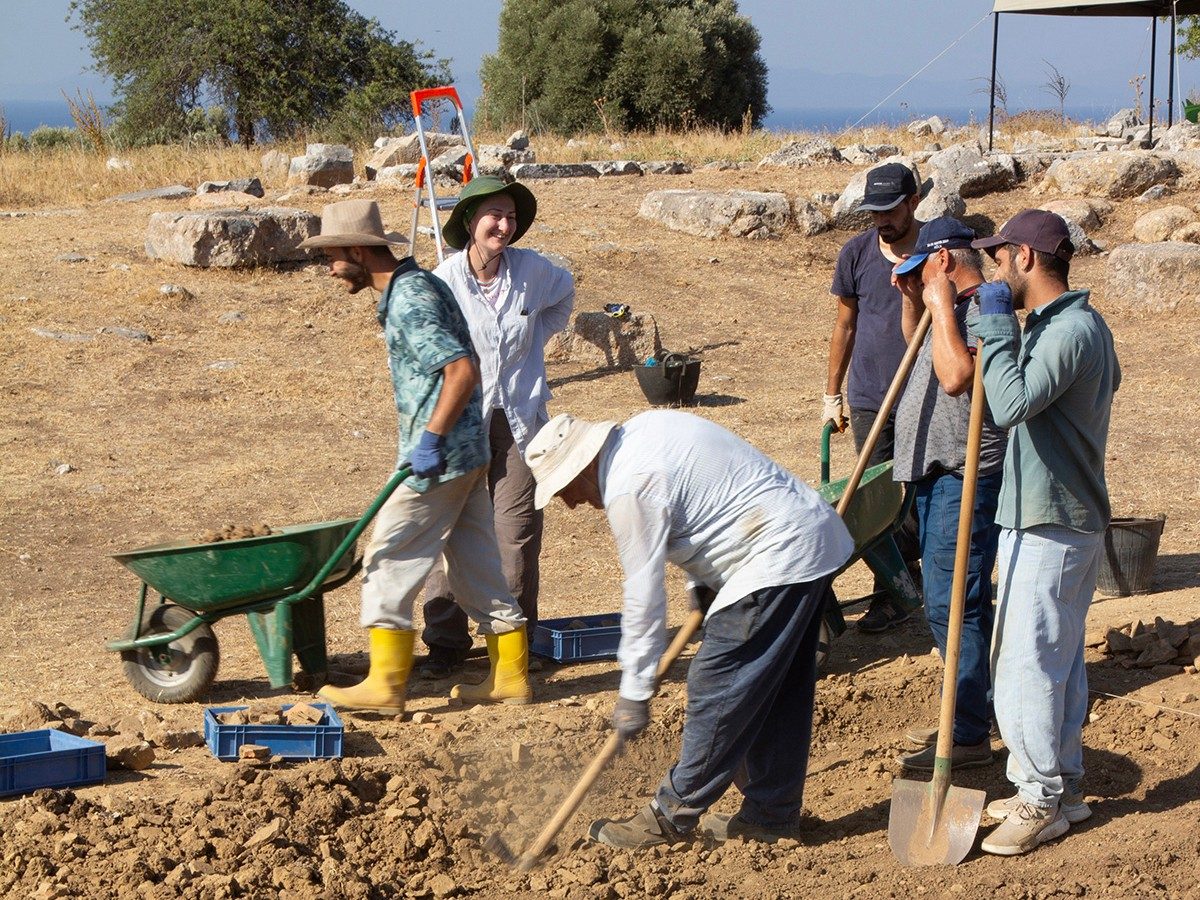 A group of six people (one woman and six men) at the archaeological site of Notion. Some handle wheelbarrows, and others stand and dig with shovels.