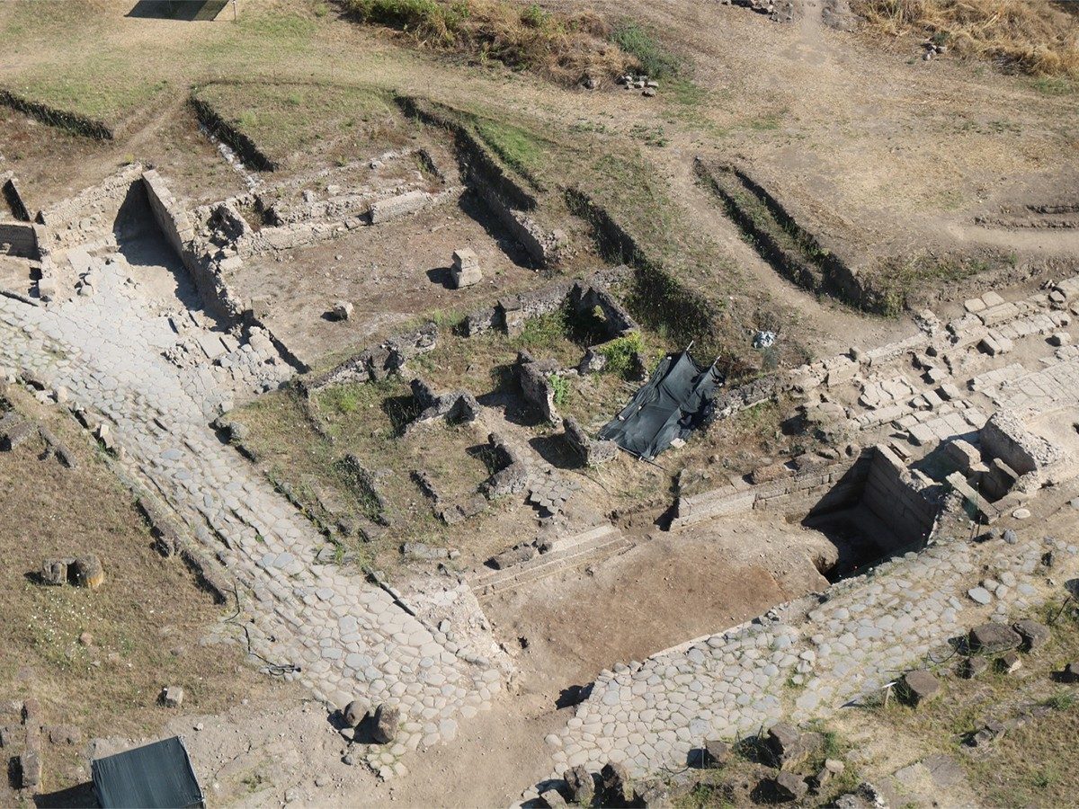 Aerial view of the archaeological site of Gabii. A stone street winds across the bottom of the image, while the foundations of several buildings can be seen rising from the grass and dirt.