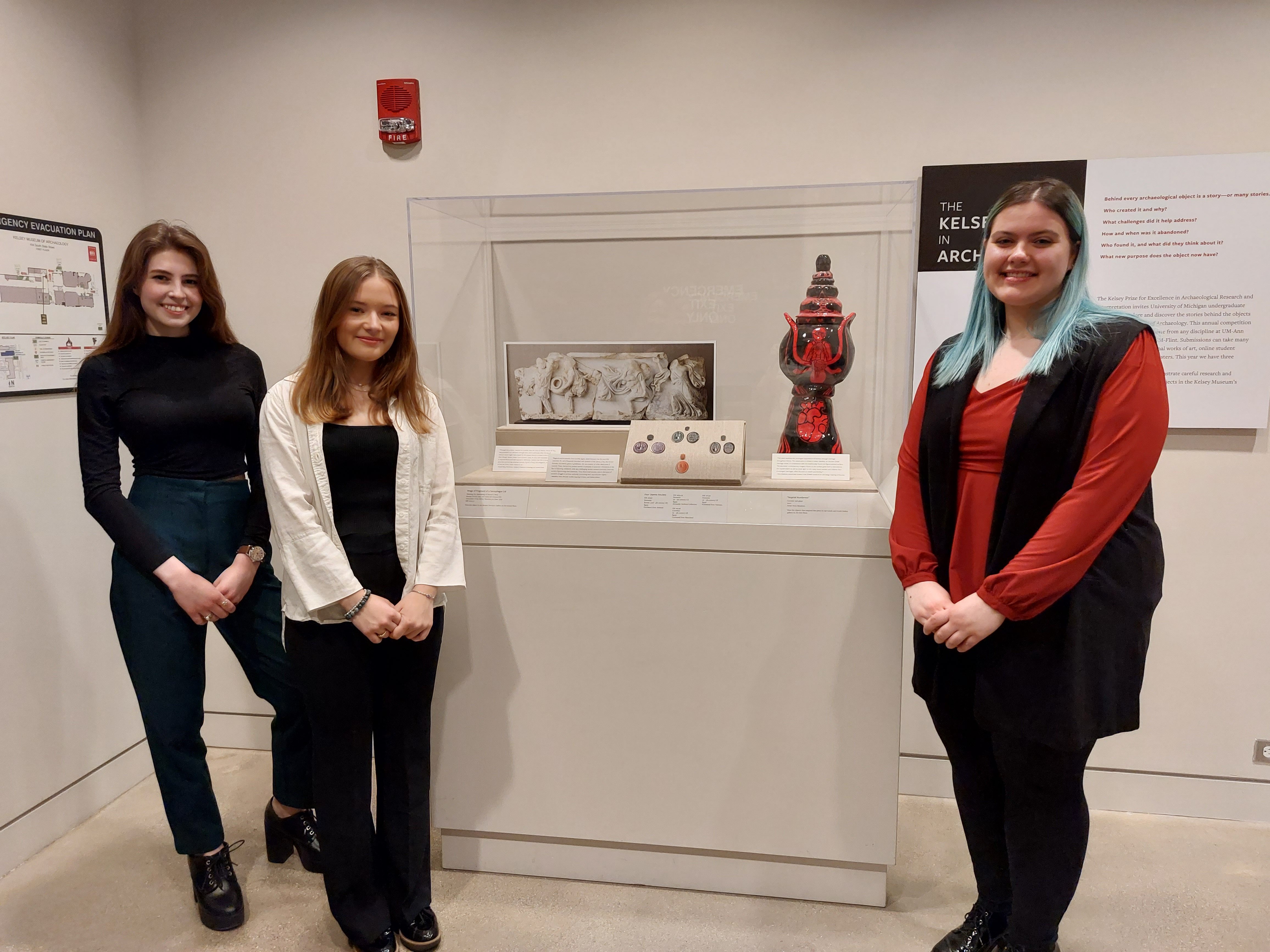 Nora Meadows, Araceli Rizzo, and Madeleine Wren stand in front of a museum vitrine containing a photo of a marble sarcophagus, photos of uterine amulets, and a black and red modern art piece inspired by an ancient Greek amphora.