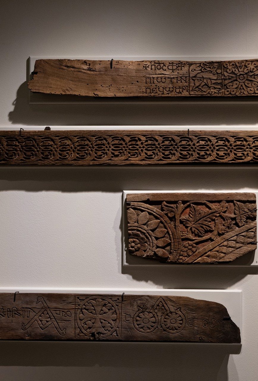 View of four long wooden lintels mounted in the Kelsey Museum. Each panel features carvings of Coptic and Byzantine imagery, including Greek letters, floral motifs, geometric shapes, and animals.
