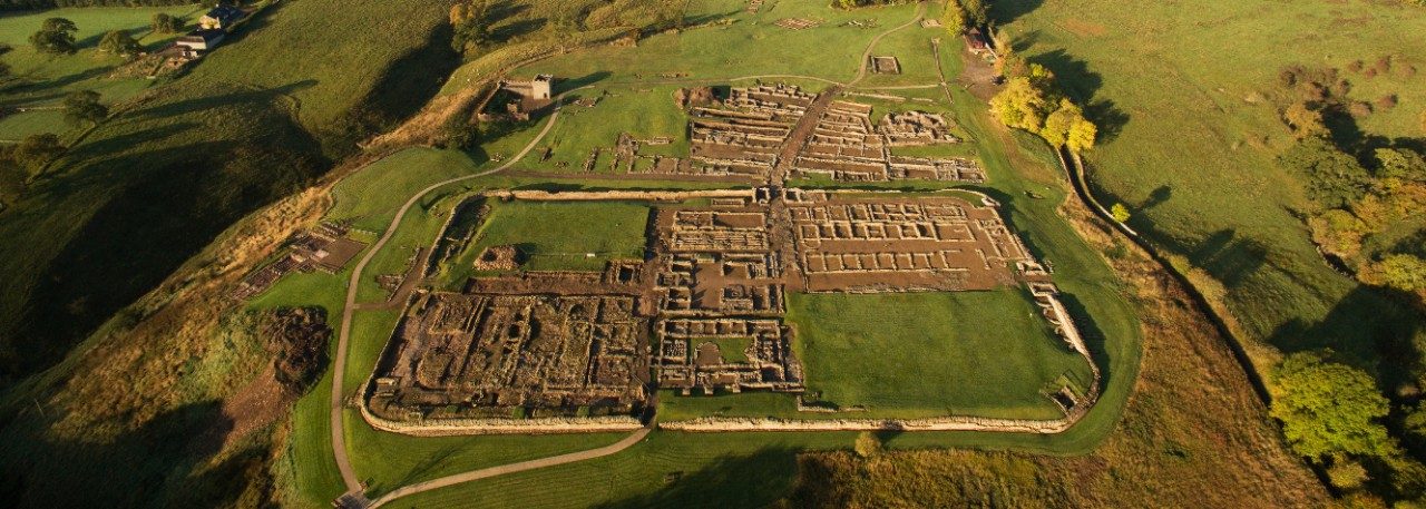 Aerial view of the archaeological site of Vindolanda in northern England. In the foreground is a fort enclosed by a stone wall, while rolling green fields are visible in the background.