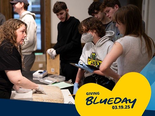 Kelsey Museum staff member Shannon Ness, wearing gloves, points to a marble inscription and talks to a group of students holding clipboards. An overlay across the image reads, “Giving Blueday, 03.19.25.”
