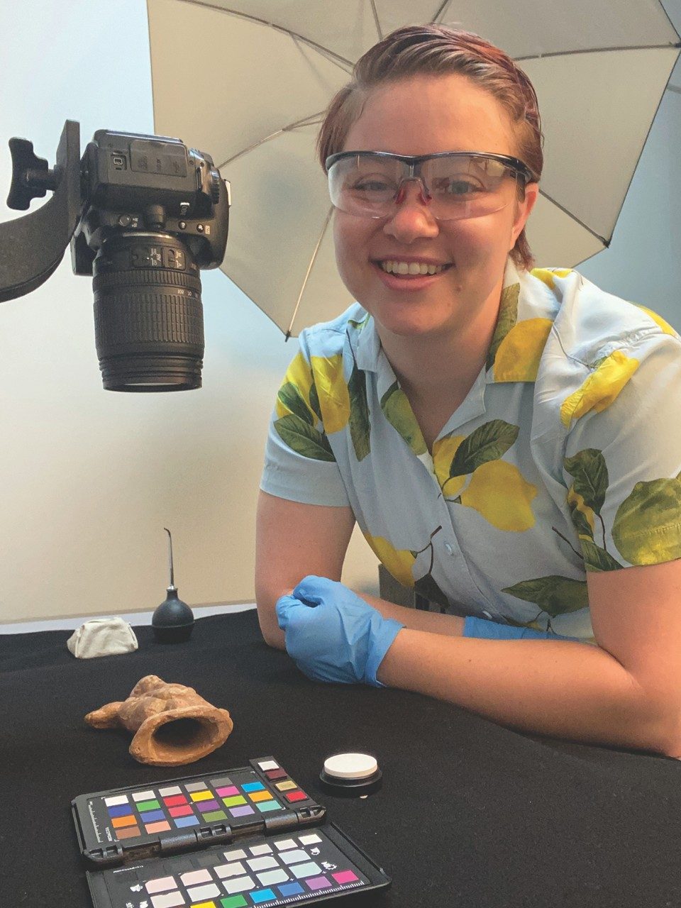 Laurel Fricker, wearing a shirt patterned with lemons, smiles from next to a photography setup in the Conservation Lab. A camera is trained on a terracotta figurine of the god Harpocrates.