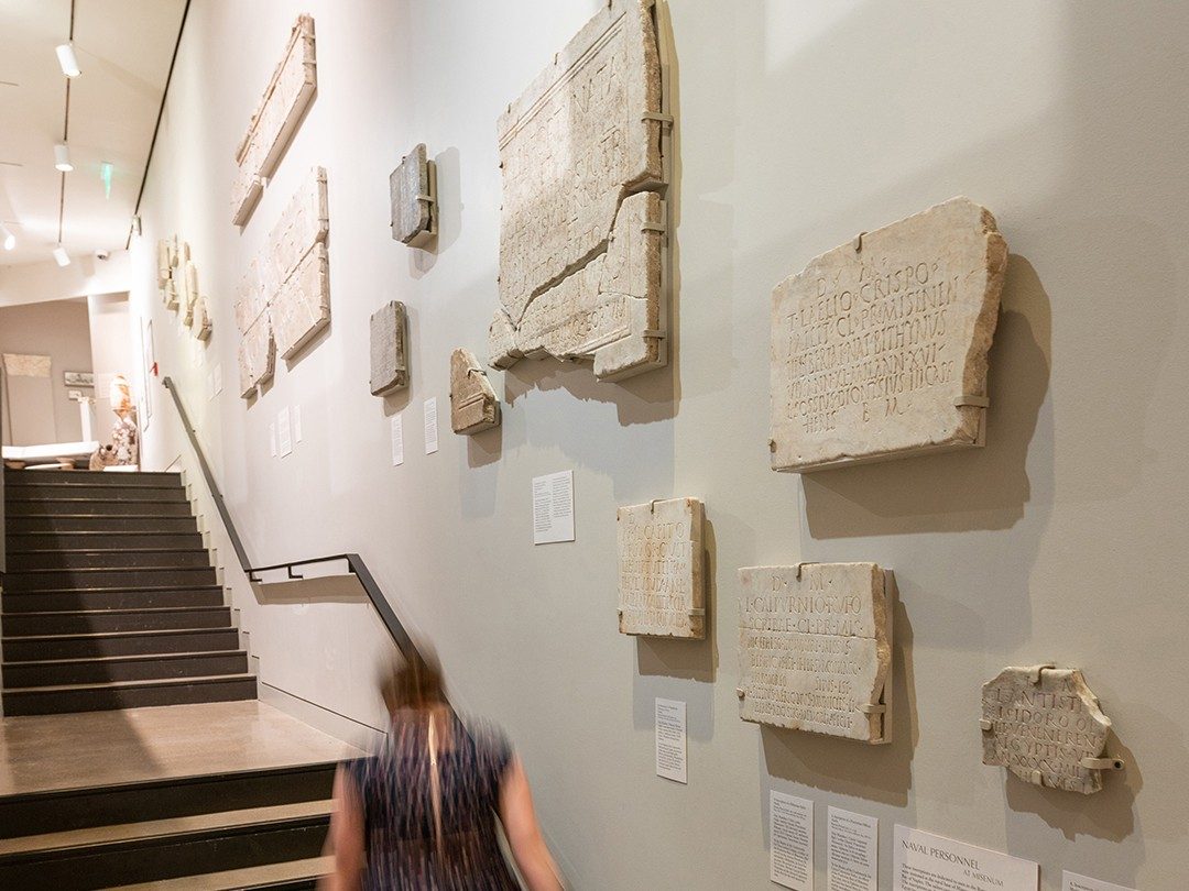 View of the stairway in the Kelsey Museum, showing a person going up the steps. The wall to the right has several white marble slabs with Latin inscriptions.