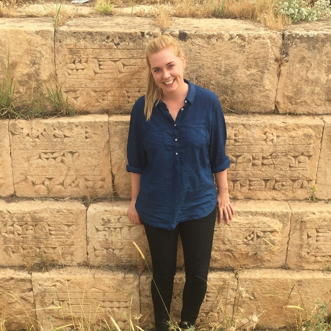Katherine Burge stands against a wall of large, stacked, stone blocks with carvings on their fronts.