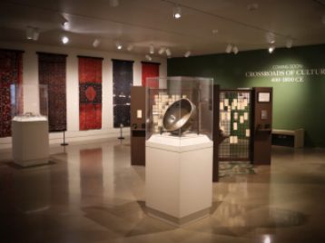 View of the Kelsey Museum galleries showing three vitrines with artifacts, the visitor feedback structure, and large-scale hanging reproductions of textiles. Text on the wall reads, “Coming Soon: Crossroads of Cultures, 400–1800 CE.”