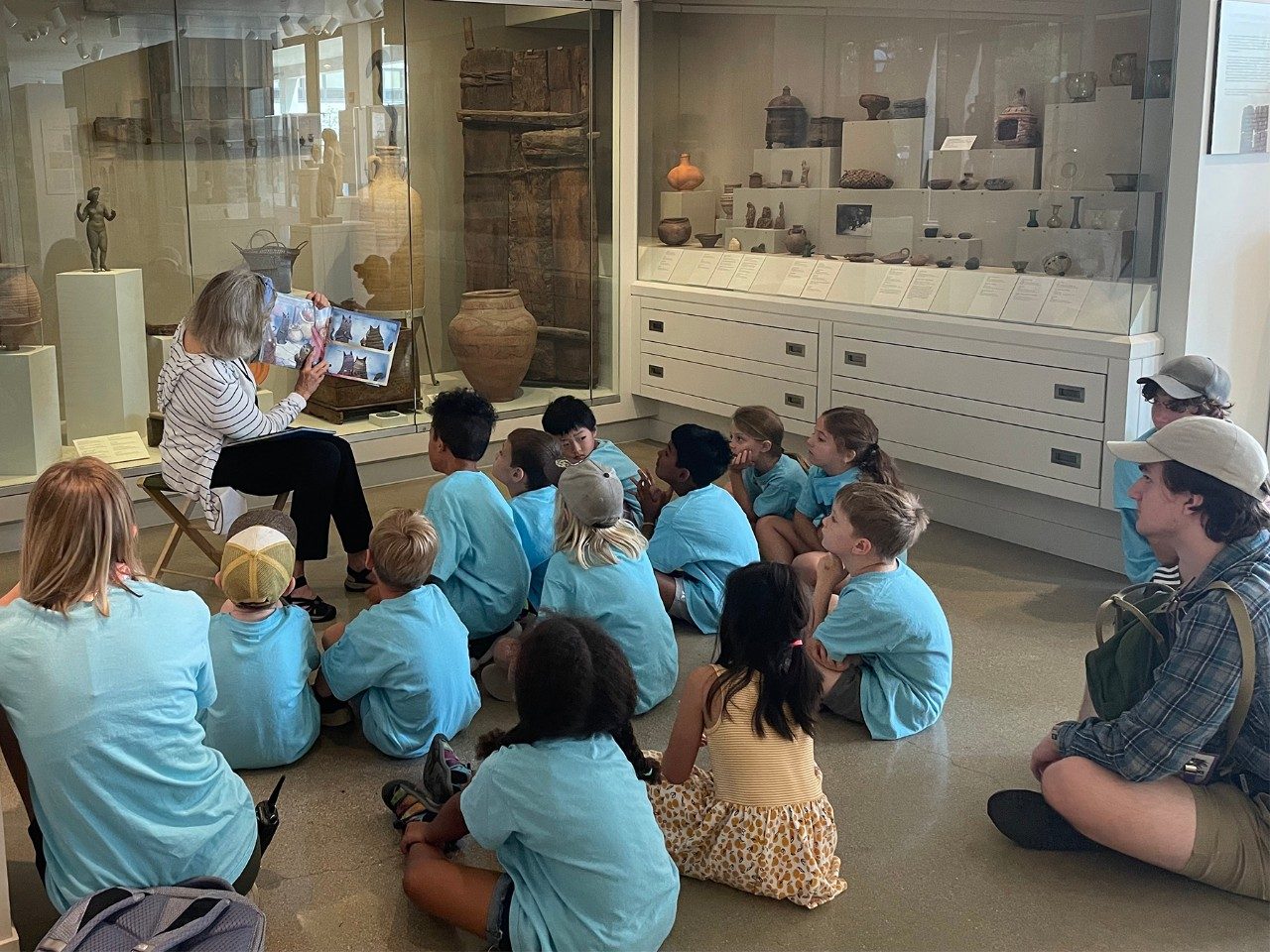 A woman holds a book up for a group of children and chaperones sitting on the floor in the Kelsey Museum.