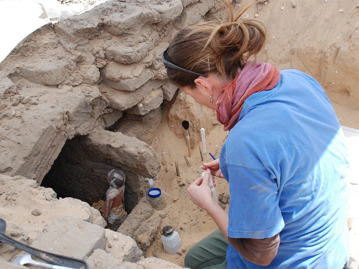 A conservator sits near the small chamber at the Abydos Middle Cemetery containing the wooden statues. She uses an array of tools and supplies, including a brush, tweezers, and tape, to temporarily repair one of the statues so it can be safely lifted.