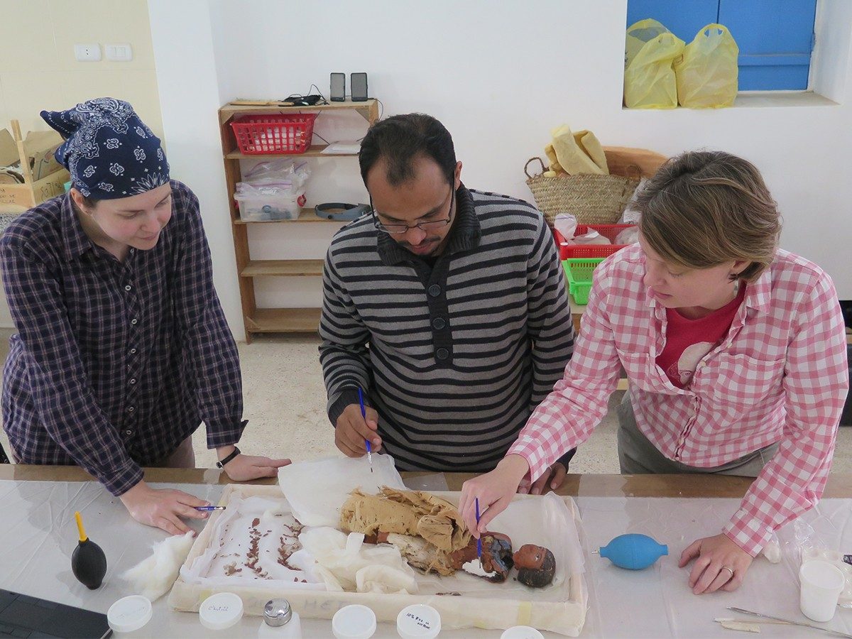 Three conservators (Carrie Roberts, Hamada Sadek, and Suzanne Davis) work on the wooden statue of the striding man. The figurine lies in a shallow box, while each of the conservators each hold a small brush.