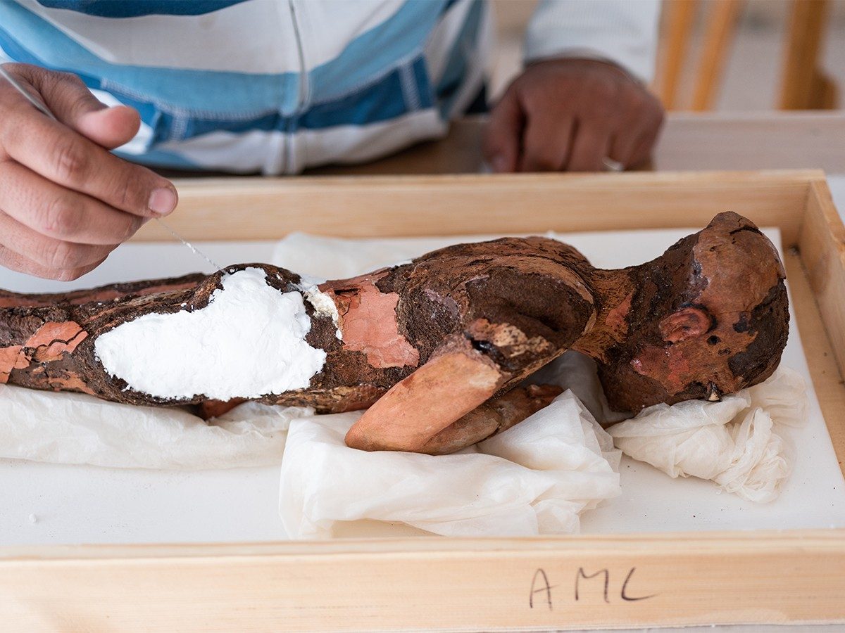Conservator Hamada Sadek works to address missing pieces of the wooden child statue, which lies facedown in a shallow wooden box.