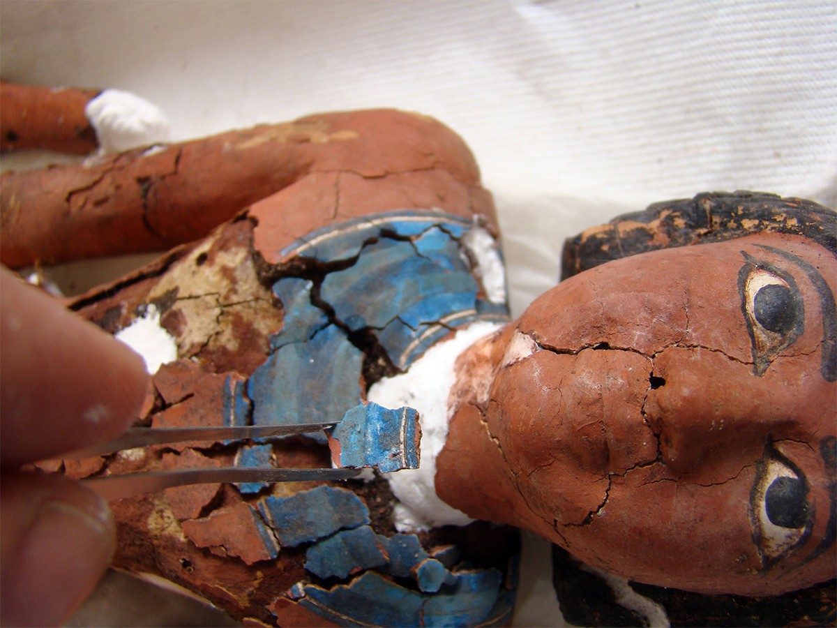 Close-up of the wooden figurine of a man. A conservator uses tweezers to carefully line up a bright blue section of the figurine’s broad collar, which had chipped away.