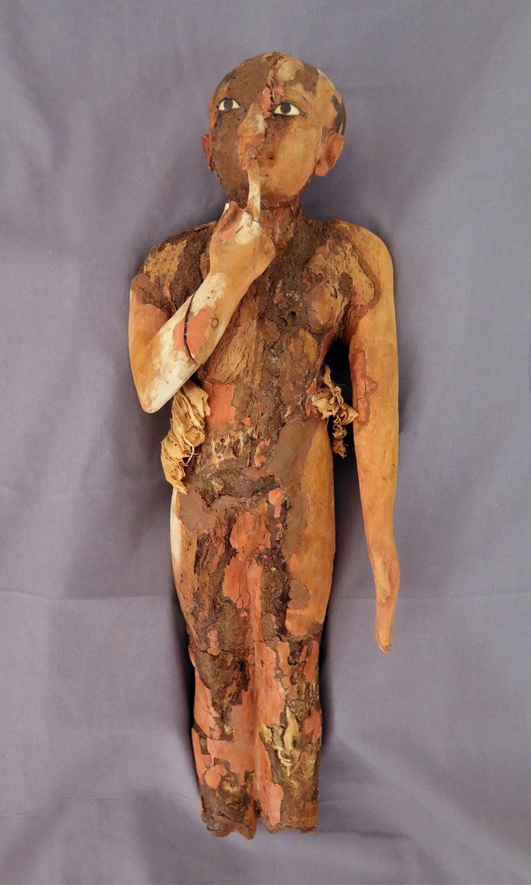 Small wooden figurine of a child, his hand upraised to his mouth, resting on a gray piece of cloth. Although the child still shows signs of wear, he has been reassembled into one piece.