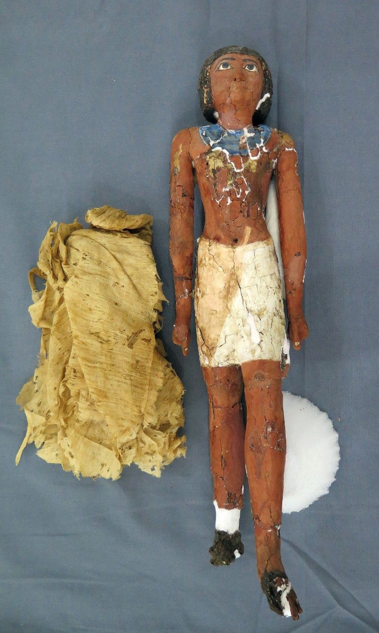 Small wooden figurine of a striding man resting on a gray piece of cloth. Although quite chipped, previously broken off pieces have been repaired. 