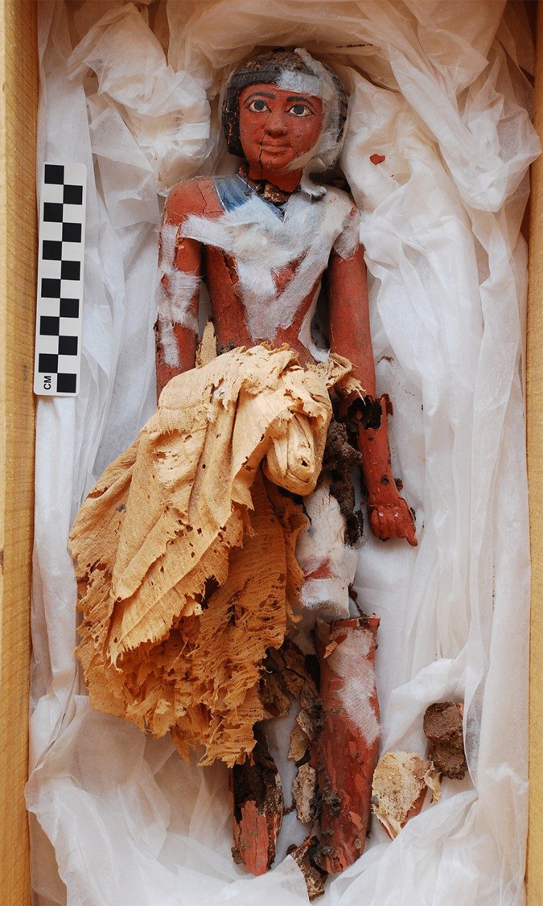 Small wooden figurine of a standing man resting in a cloth-lined box next to a measuring stick and pieces that have broken off. Thin bands of white, temporary tape hold the figurine together, and a bundle of cloth rests across his lap. 