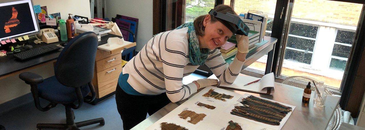 Suzanne Davis wearing a headband magnifier pushed up on her forehead, leaning over a metal table with several ancient, colorful textiles laid out flat. In the background is a desk with a computer, on which an image of a textile is displayed. 